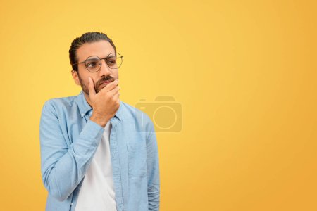 Pensive indian man in a denim shirt poses thoughtfully with hand on chin against a uniform yellow background