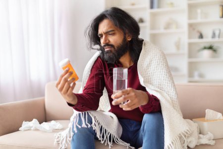 A worried Indian man wrapped in a blanket looks confusedly at a bottle of pills while holding a glass of water, illustrating illness and treatment