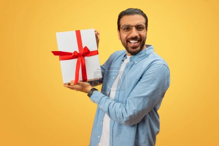 Téléchargez les photos : Homme oriental avec un grand sourire offrant une boîte cadeau blanche avec ruban rouge, s'engageant directement à la caméra - en image libre de droit