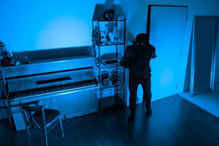 In a blue-lit room, a thief man checking for valuables, with a piano in the foreground, full length