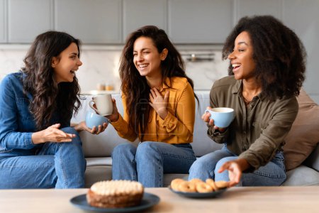 Chat animé entre trois jeunes femmes multiraciales avec café et pâtisseries sur un canapé confortable dans un salon à la maison