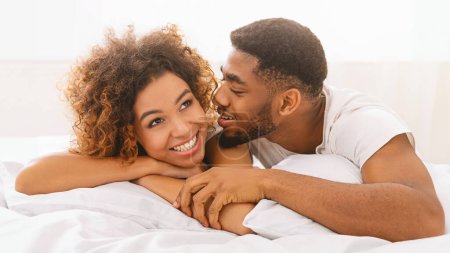 Téléchargez les photos : Amoureux afro-américains heureux couchés dans le lit à la maison, homme chuchotant secret sur son oreille de petite amie - en image libre de droit