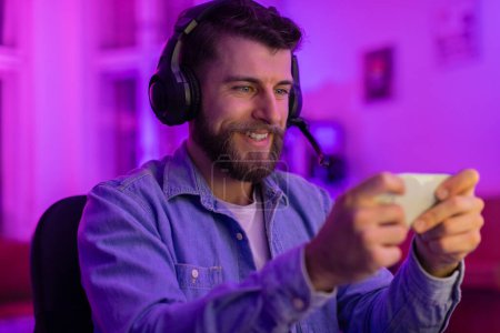 Photo for Happy young man gamer with headphones enjoying playing games on his smartphone with colorful lighting - Royalty Free Image