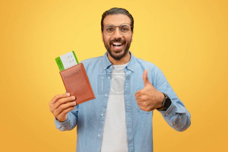Téléchargez les photos : Homme indien joyeux montre l'approbation avec passeport et cartes d'embarquement sur jaune, signalant une expérience de voyage positive - en image libre de droit