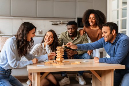 Un grupo de amigos multirraciales comprometidos participan en un tenso juego de torre de bloques de madera en una mesa, mostrando concentración y diversión