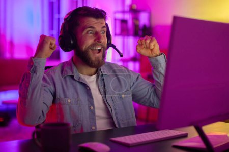 A man in a casual denim shirt with headphones raises his fists in victory or excitement while sitting in a vibrant, neon-lit gaming setup