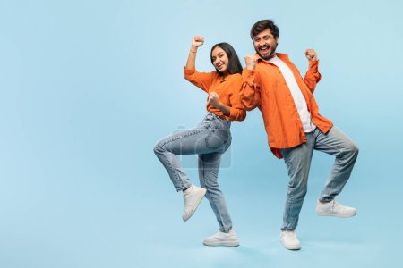 Una alegre pareja india joven con chaquetas naranjas y vaqueros bailan con los brazos en una pose alegre sobre un fondo azul
