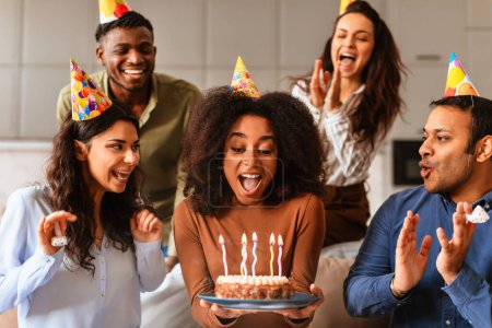 Freunde verschiedener Rassen umgeben eine Frau, die eine Geburtstagstorte in der Hand hält und in einem Moment der Aufregung gemeinsam Kerzen ausbläst