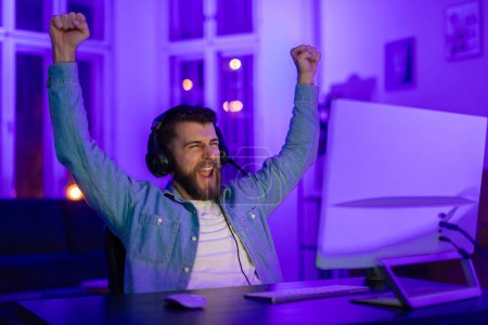 Man with headset celebrates a gaming achievement with arms raised and a joyful shout, home interior