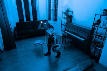 Téléchargez les photos : Homme cambrioleur avec masque sur l'exploration appartement sombre avec lampe de poche, tenant sac à dos, vue grand angle - en image libre de droit