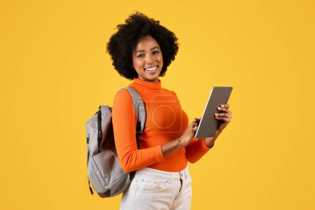 Photo for Confident smiling African American woman student with curly hair smiling and holding a digital tablet, with a backpack slung over her shoulder, against a yellow background - Royalty Free Image