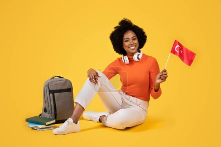 Photo for Joyous woman with headphones around her neck, holding the Turkish flag, sitting with books and a grey backpack, wearing a cozy orange turtleneck and white pants, against a yellow background - Royalty Free Image