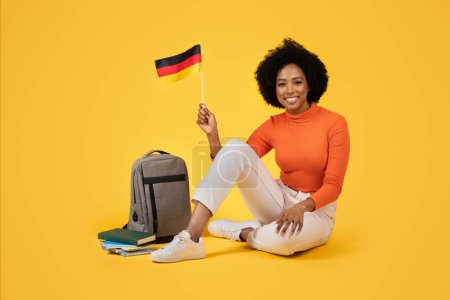 Foto de Mujer joven y feliz sosteniendo orgullosamente la bandera alemana, sentada con las piernas cruzadas junto a su mochila gris y libros, con un cuello alto naranja ajustado y pantalones blancos sobre un fondo amarillo - Imagen libre de derechos
