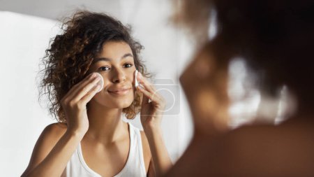 Téléchargez les photos : Belle fille afro-américaine nettoyant visage avec des tampons de coton regardant dans le miroir à la salle de bain. Concept de démaquillage quotidien - en image libre de droit