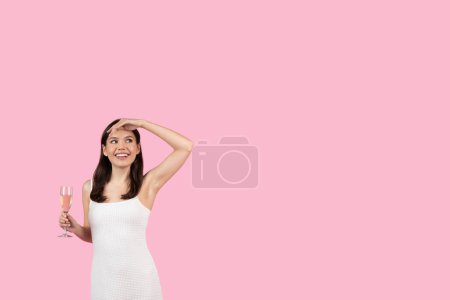 Téléchargez les photos : Une femme souriante en robe blanche tient une coupe de champagne, regardant au loin sur un fond rose - en image libre de droit