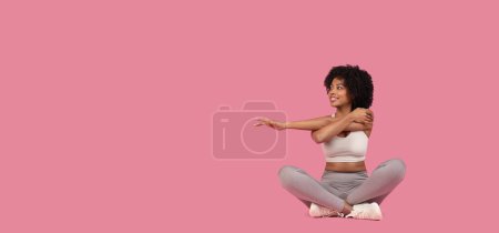 Smiling young african american woman in gray sportswear sits cross-legged, stretching her arms on a pink background