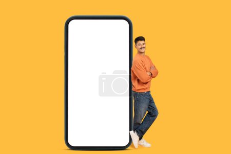Un homme souriant et debout avec les bras croisés à côté d'un smartphone géant avec écran blanc, adapté aux utilisations maquettes