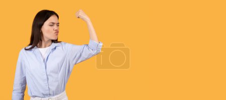 Photo for Confident woman flexes her bicep, making a strong pose, with a determined look on a yellow backdrop, panorama with copy space - Royalty Free Image