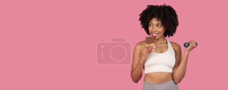 Photo for African american woman balances indulgence with fitness by playfully eating chocolate while holding a dumbbell - Royalty Free Image
