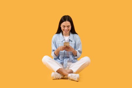 Young pretty woman in casual clothes using her phone, sitting cross-legged against a yellow background
