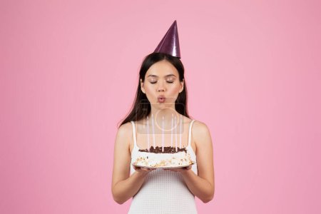 Una mujer joven con un sombrero de fiesta sopla velas en un pequeño pastel de cumpleaños, celebrando con un fondo rosa