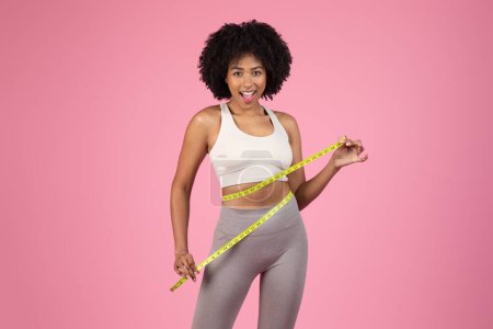 Foto de Emocionado joven afroamericana mujer en traje de fitness felizmente mostrando su delgada cintura con una cinta métrica - Imagen libre de derechos