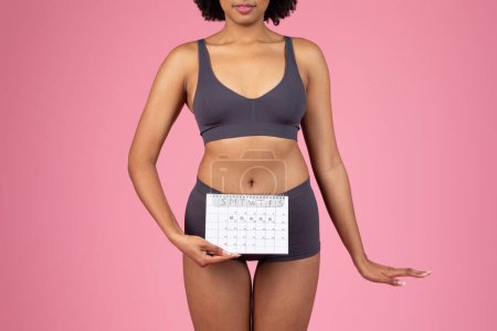 A young African American woman in a sports bra is holding a menstrual cycle calendar against her lower abdomen on a pink backdrop
