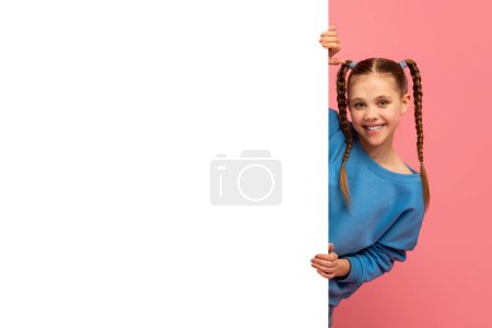 Téléchargez les photos : Jeune fille souriante aux cheveux tressés regardant par derrière un tableau blanc vierge sur fond rose - en image libre de droit