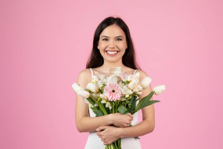 Photo for A radiant young pretty woman in a white dress tenderly embraces a bouquet, against a pink-hued backdrop - Royalty Free Image