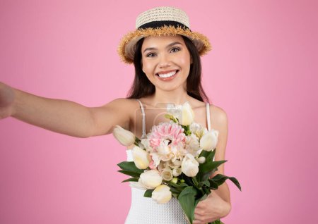 Photo for Excited woman in a straw hat takes a selfie with a bouquet of flowers, against a pink studio background - Royalty Free Image