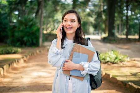 Téléchargez les photos : Joyeux jeune femme communique sur un téléphone intelligent tenant des ordinateurs portables dans la scène extérieure au parc public - en image libre de droit