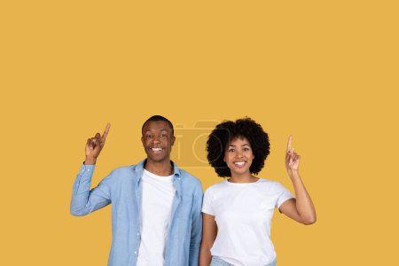 Téléchargez les photos : Jeune couple joyeux afro-américain pointant vers le haut, indiquant des idées ou des aspirations dans un contexte jaune - en image libre de droit