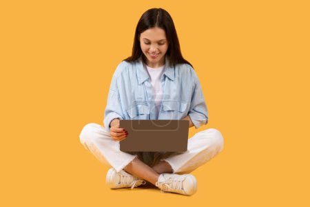 A focused woman engaged with a laptop while comfortably seated on the floor on a yellow background