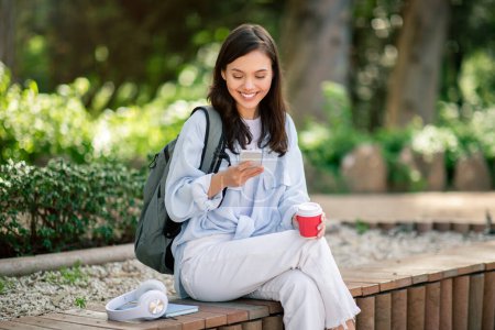Una joven estudiante relajada con mochila comprueba su teléfono inteligente y disfruta de un café durante una visita al parque