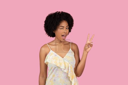 Foto de Mujer afroamericana en vestido de verano mostrando signo de paz con una cara juguetona sorprendida sobre fondo rosa - Imagen libre de derechos