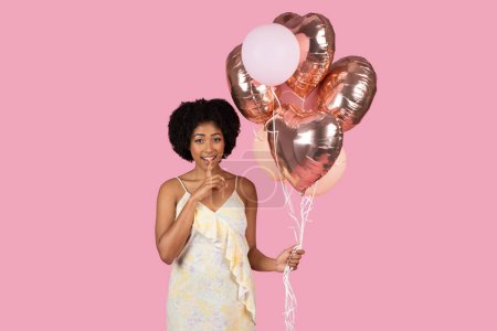 Téléchargez les photos : Une femme afro-américaine souriante tient des ballons en forme de cœur en or rose, suggérant une célébration ou une occasion spéciale - en image libre de droit