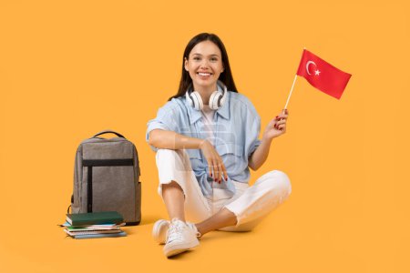 Une jeune femme souriante arborant un drapeau de Turquie, avec écouteurs et fournitures scolaires sur le côté sur fond jaune