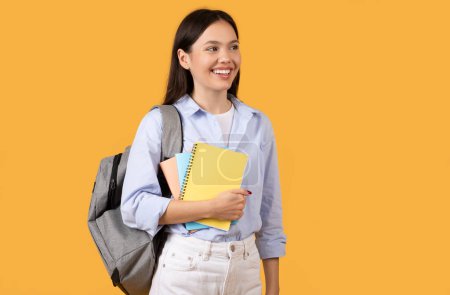 Photo for A cheerful young woman holding notebooks and looking to the side on a yellow backdrop, copy space - Royalty Free Image