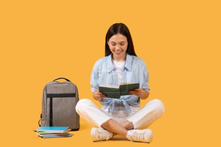 Young brunette lady enjoying a book while sitting cross-legged on a yellow background with school supplies