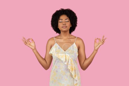 Heitere Afroamerikanerin meditiert in friedlicher Pose mit einem leichten floralen Kleid vor rosa Hintergrund