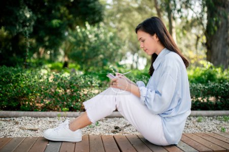 Una joven mujer se sienta en el suelo en un entorno de parque sereno, comprometida con su teléfono inteligente, rodeada de exuberante vegetación