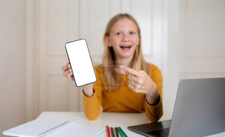Foto de Una chica adolescente alegre apuntando a la pantalla del teléfono inteligente en blanco con una expresión alegre, un niño adolescente feliz sentado en un escritorio con una computadora portátil, estudio en casa, recomendando una nueva aplicación, maqueta - Imagen libre de derechos