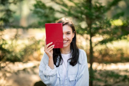 Mujer en un parque sosteniendo un libro rojo cubriéndose parcialmente la cara, sonriendo a la cámara, jovencita disfrutando leyendo