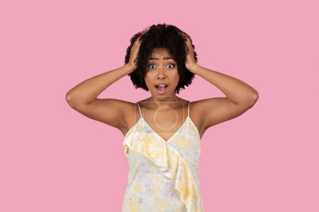 Téléchargez les photos : Surprise jeune femme afro-américaine dans une robe d'été avec ses mains sur la tête, expression large sur un fond rose - en image libre de droit