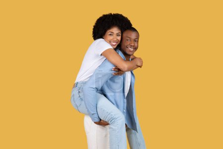 Verspieltes und liebevolles afroamerikanisches Paar, wo die Frau huckepack auf dem Rücken eines Mannes vor gelbem Hintergrund reitet