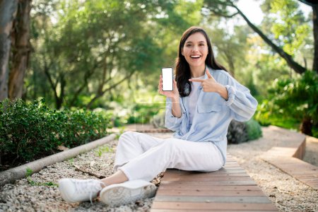 Foto de Mujer con ropa casual sentada en el camino de madera que muestra una pantalla de teléfono inteligente en blanco, árboles en el fondo - Imagen libre de derechos