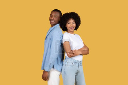 Zwei lächelnde junge afroamerikanische Paare stehen Rücken an Rücken, posieren bequem vor gelbem Hintergrund und strahlen Zuversicht und Zufriedenheit aus.