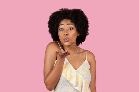 Enchanting African American woman sending a flying kiss with her hands, cute and playful on a pink backdrop