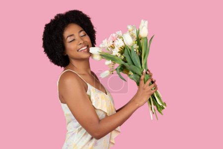 Téléchargez les photos : Une joyeuse Afro-Américaine aux cheveux bouclés étreint doucement un bouquet de tulipes blanches et autres fleurs, les yeux fermés dans le délice, sur un fond rose - en image libre de droit