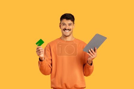 Man smiling and displaying a credit card and tablet, signifying online shopping, isolated on a yellow background
